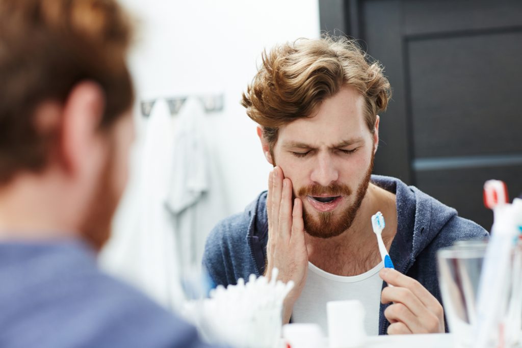 Image of a man holding cheek in pain while brushing teeth.