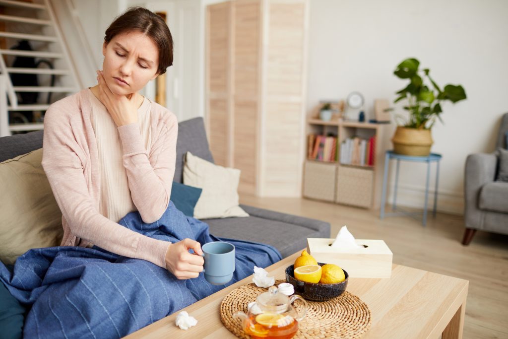 A person sitting down with a blanket and mug, holding their throat. Used tissues are scattered on a coffee table.