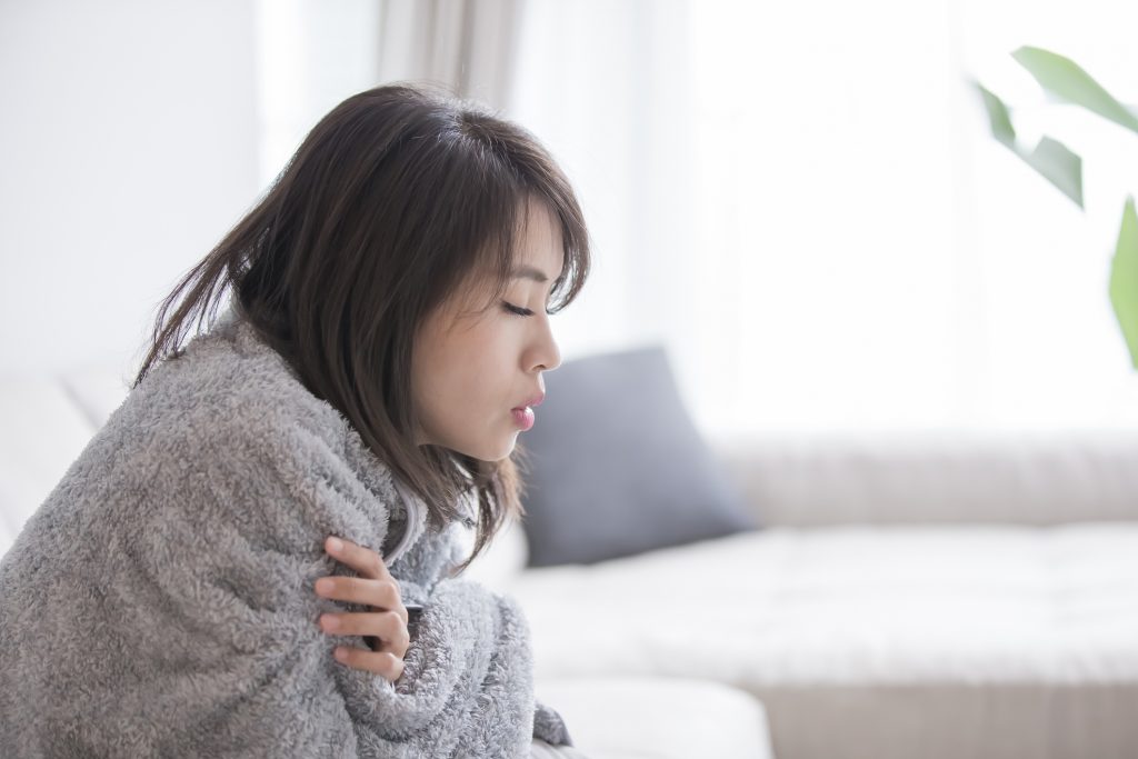 A woman sits on a cough with blanket wrapped around her shoulders, eyes closed and exhaling.