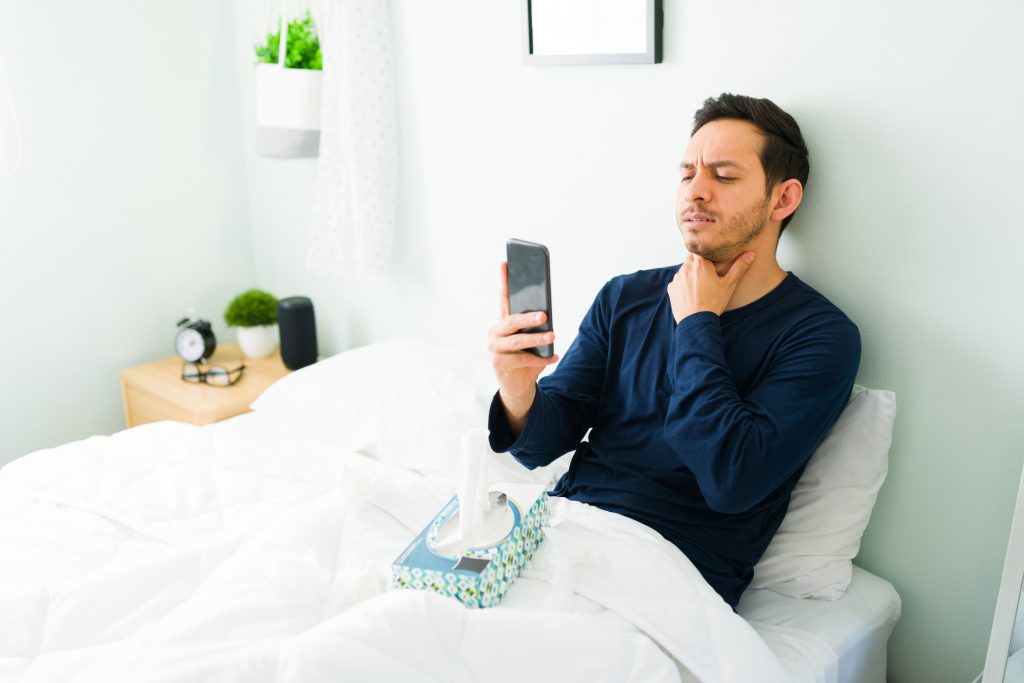 A person sitting up in bed, holding throat and video calling with someone on their phone. A box of tissues sits on the bed.