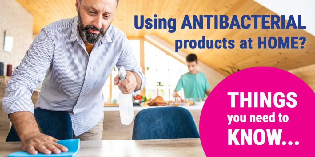 Image of man cleaning dining table with spray bottle and cloth. In the background, a teenager prepares food in the kitchen. Overlaid text reads: "Using antibacterial products at home? Things you need to know."